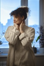 Young woman looks out the window while touching her face in a cozy indoor setting during daytime