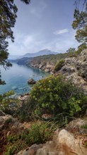 Rocky coastal landscape with clear water, lush vegetation and a Tahtali mountain in the background,