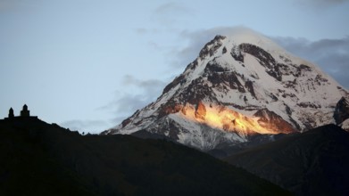 Sunbeams illuminate a mountain peak in the evening light with church in the foreground, Gergeti