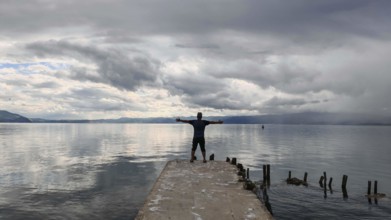 Person standing with outstretched arms on a pier surrounded by water and clouds, Lake Ohrid, North