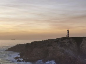 Landscape with lighthouse on rocks, illuminated by sunset, with calm sea and clouds in the