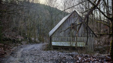Snowy trail leads to a secluded wooden hut in the winter forest in front of a range of hills,