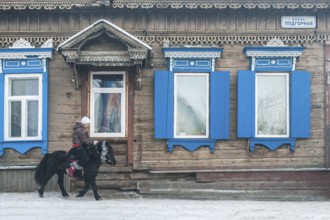 Irkutsk, Siberia, Russia. A young woman riding a pony in front of traditional wooden houses during
