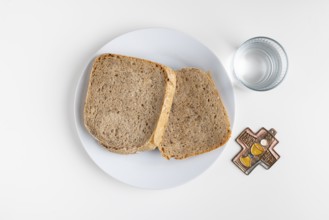 Two slices of bread on a plate with water glass and cross symbol on a plain white background, Lent