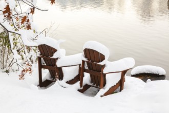 Two brown stained wooden Adirondack chairs by riverside covered with winter like snow in late