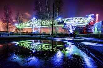 Intensely illuminated bridge construction at night with reflection in water and dramatic clouds,