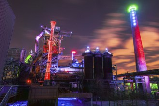 Night view of an illuminated industrial plant with colored lights and clouds in the sky,