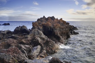 Rough rock formation jutting into the sea surrounded by waves under a slightly cloudy sky, Puerto