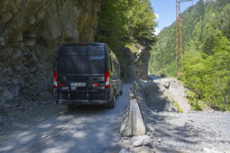A black van drives on a narrow mountain road surrounded by rocks and thick vegetation, camper