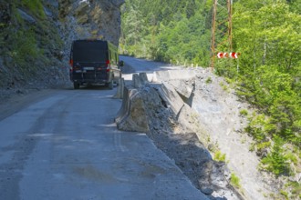 A black van drives on a remote road along a steep cliff with green vegetation, camper drives on