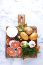 Potatoes, salmon, garlic, onion, and herbs are placed on a wooden cutting board. These ingredients