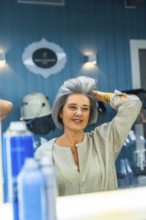 Mature woman looking at her reflection, touching her fresh gray hairstyle, and smiling in a modern