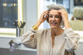 Mature woman looking at her reflection in the mirror, touching her styled gray hair and smiling,