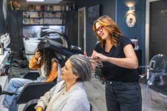 Hairstylist smiling while prepping a senior woman's hair for treatment as another client dries