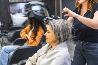 Hair stylist blow drying gray hair of a senior woman sitting in a hair salon, with another client