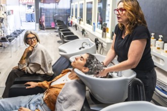 Hairdresser shampooing a young woman at a modern salon while an older client watches nearby,