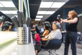 Hairbrushes standing ready on a white counter as a stylist blow dries a senior woman's hair in a