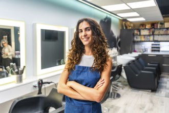 Confident young woman with curly hair wearing an apron and smiling, standing with crossed arms in