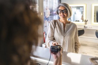 Senior woman smiling while making a digital payment with a credit card at a salon's reception desk,
