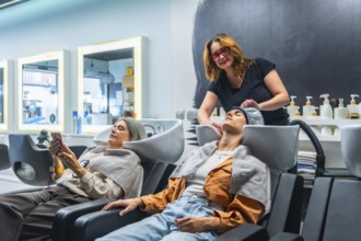 Hairdresser washing young woman's hair at a sleek salon basin while another client relaxes on her