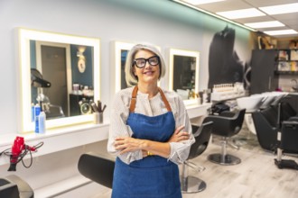 Senior woman hairstylist smiling, standing with arms crossed, wearing glasses and an apron,