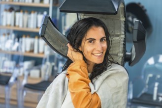 Woman relaxes under a salon hair dryer during a pampering hair treatment, smiling with wet hair as