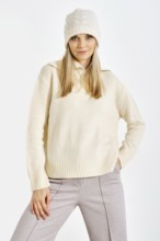 A model stands confidently in a studio while wearing a light sweater and a knitted hat. The setting