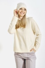 A woman stands smiling while wearing a light sweater and a warm hat. She has long blonde hair and