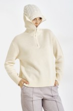 A woman stands in a neutral background wearing a warm knitted sweater and a hat. She looks relaxed