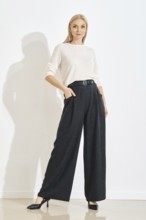 A woman in grey pants and a white top poses in a simple indoor space. Her hands are in her pockets.