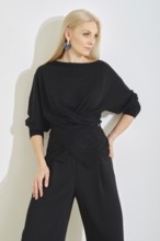 A woman poses gracefully in a modern indoor space, wearing a black wrap top and wide-leg pants. Her