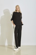 A woman stands confidently in a stylish black tracksuit featuring white stripes along the sides.