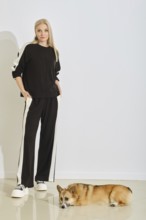 A woman stands in a sporty black tracksuit with white stripes while her dog lies comfortably on the