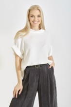 A woman with long blonde hair is smiling while standing indoors. She wears a casual white top and