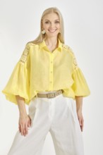 Cheerful woman in a vibrant yellow blouse with lace details and white trousers posing in bright