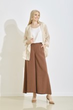 A smiling woman stands gracefully in a bright, minimalist studio, dressed in a light top and