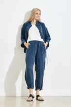A woman is standing against a white wall in a bright room. She wears a blue pants and jacket with a