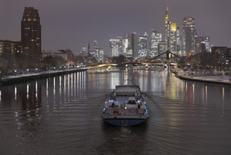 A ship sails towards the snow-covered and glowing Frankfurt banking skyline in the evening,