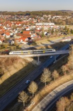A train on a bridge crosses a country road surrounded by houses and trees in a rural area, test run