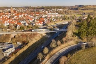 Train crosses a bridge in a town with red roofs and surrounding forest, test run on the new