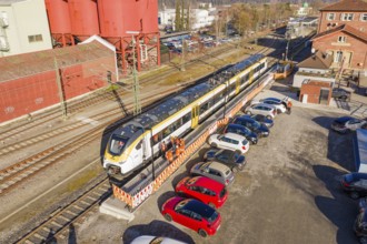 Train at train station next to red buildings with parked cars and people working in orange vests,