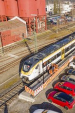 Close-up of a train at a station surrounded by industrial buildings, cars and several workers, test