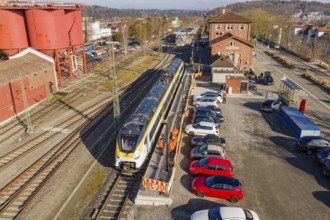Train stands at a train station next to parked cars and industrial buildings, several workers in