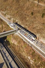 Train travels through hilly landscape over bridge surrounded by vegetation and tracks, test run on