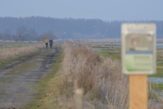 Two people ignore a protected area sign and walk along a blocked dirt road, surrounded by nature in