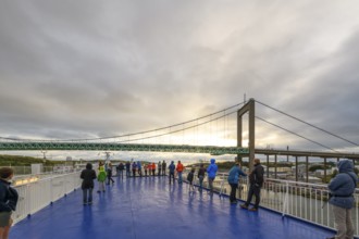 People on a blue deck looking at a bridge and dramatic sky