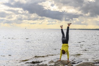 Young woman girl doing a handstand on rocky archipelago at Lake Vänern under cloudy sky, Värmlands