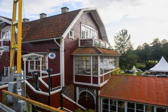 Two-storey red house with glass veranda against wooded background under blue, Sjötorp, Västra