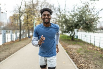 Young black man running along a path by the river, smiling at the camera and engaging in fitness