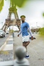 Young black man in sportswear stretching before a workout, standing on a sidewalk with the eiffel